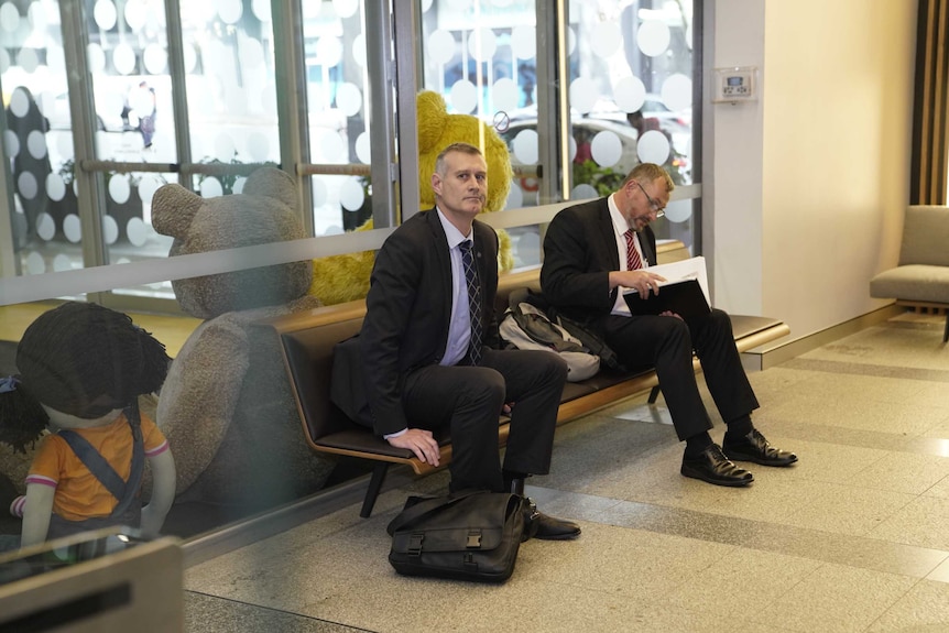 Two men in suits wait on a bench seat.