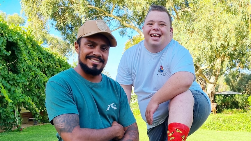 kyran and bill stand on a field of green grass with a clear blue sky above, both smiling. bill has his foot on kyran's leg
