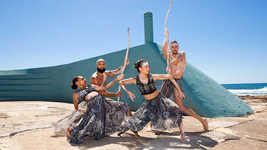 4 dancers, two women & two men, dressed in black &  white and wearing ochre, in front of turquoise wall with ocean in back, sun.