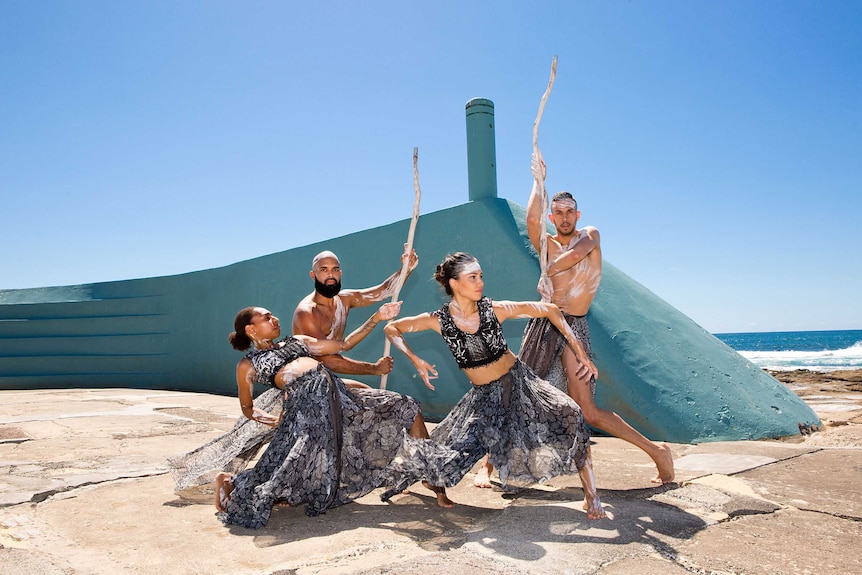 4 dancers, two women & two men, dressed in black &  white and wearing ochre, in front of turquoise wall with ocean in back, sun.