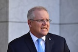 Scott Morrison looks to the right as he stands at a podium mid-speech. He wears a light blue tie, a dark blue suit and glasses.