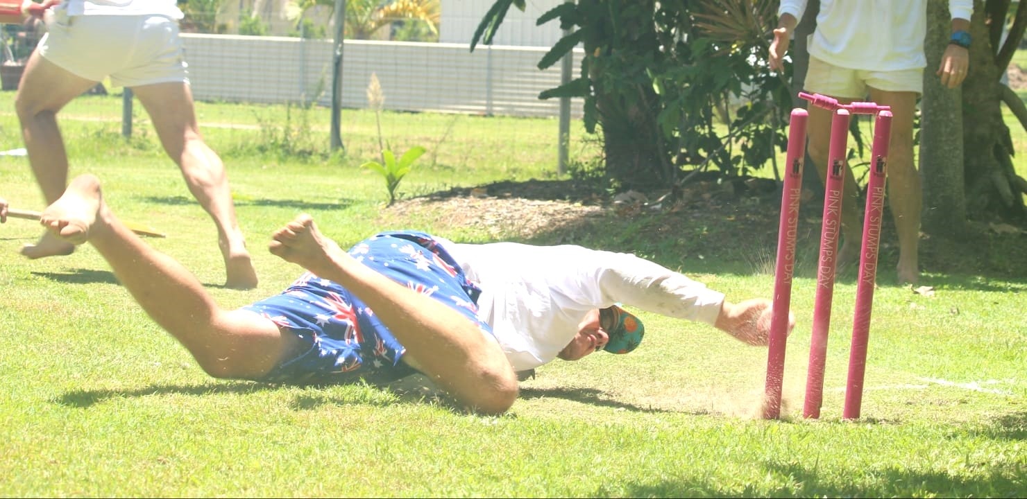 A man wearing a long sleeved white cricket shirt and blue board shirts dives towards pink cricket wickets.