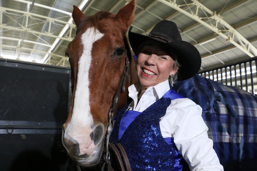 A woman in a black hat and blue sequined top stands beside a horse.