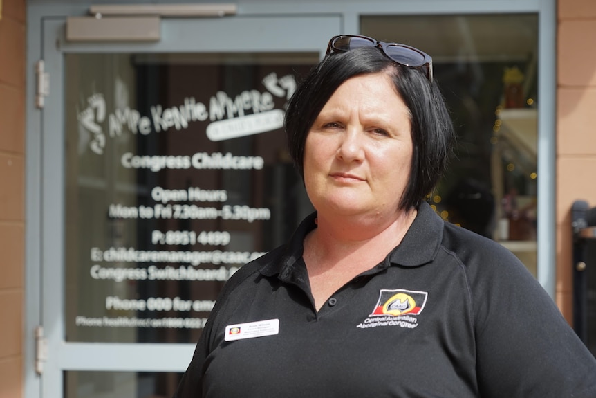 A woman in a black polo shirt looks seriously at the camera. She is in front of a daycare centre.