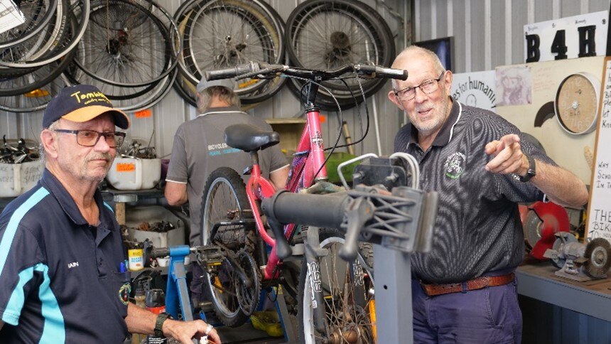Two men stan either side of a bike on stand. One is pointing in the distance. 