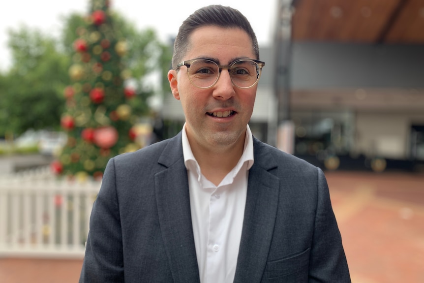 A profile of a man looking ahead wearing a dark blazer, white shirt and glasses.