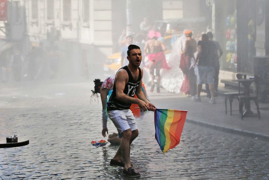 A young man holding a rainbow flag stands in the street shouting.