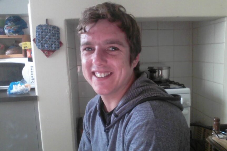 A man dressed in a grey hoody sits in a kitchen and smiles toward the camera
