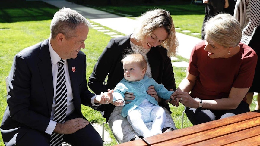 A baby in a blue knitted jacket looks at Bill Shorten (left) as mother Alicia Payne (centre) and Tanya Plibersek (right) smile