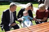 A baby in a blue knitted jacket looks at Bill Shorten (left) as mother Alicia Payne (centre) and Tanya Plibersek (right) smile