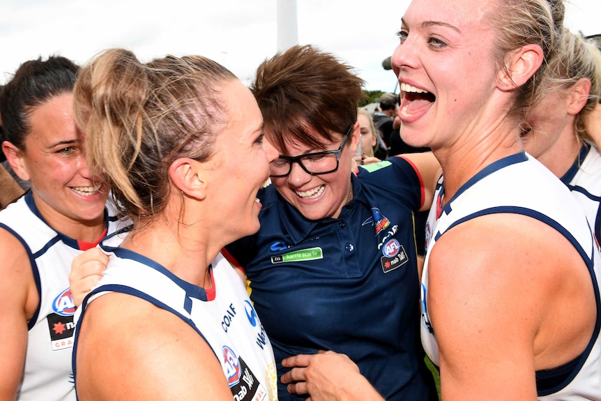 Bec Goddard smiles as she is surrounded by celebrating Adelaide Crows players in a changeroom