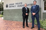Two middle aged men with dark suits and ID badges stand outside police station with arms in front.