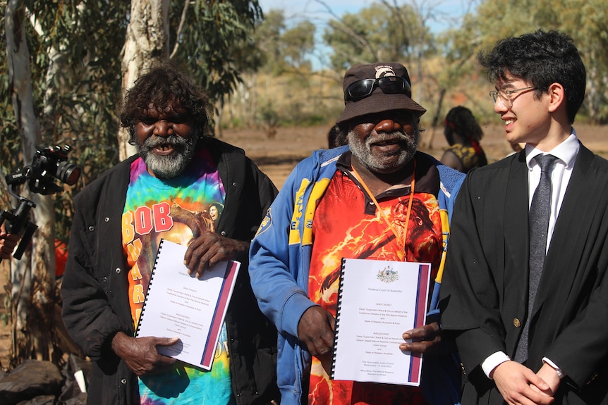 Two Aboriginal men in colourful clothes stand next to a man in black robes holding up a document and pointing to it