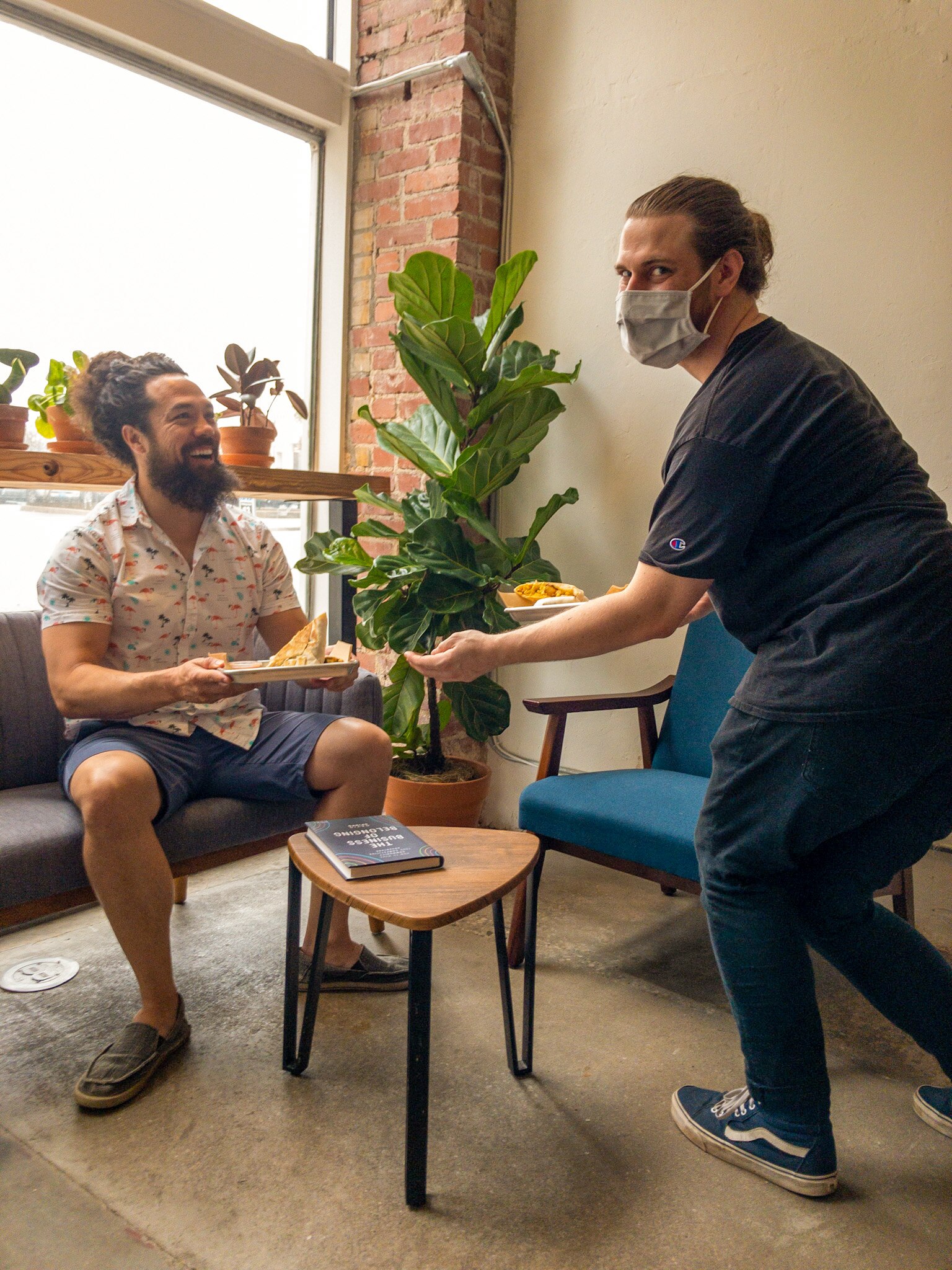 A seated man grins as another man in a face mask hands him food in a cafe