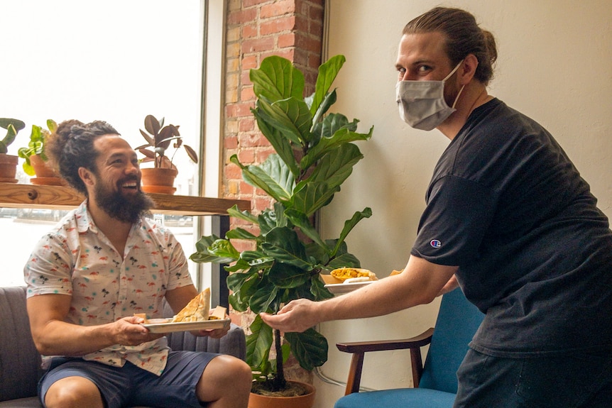 A seated man grins as another man in a face mask hands him food in a cafe