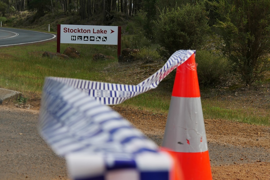 Police tape strung between two witches hats blocking access to a gravel road