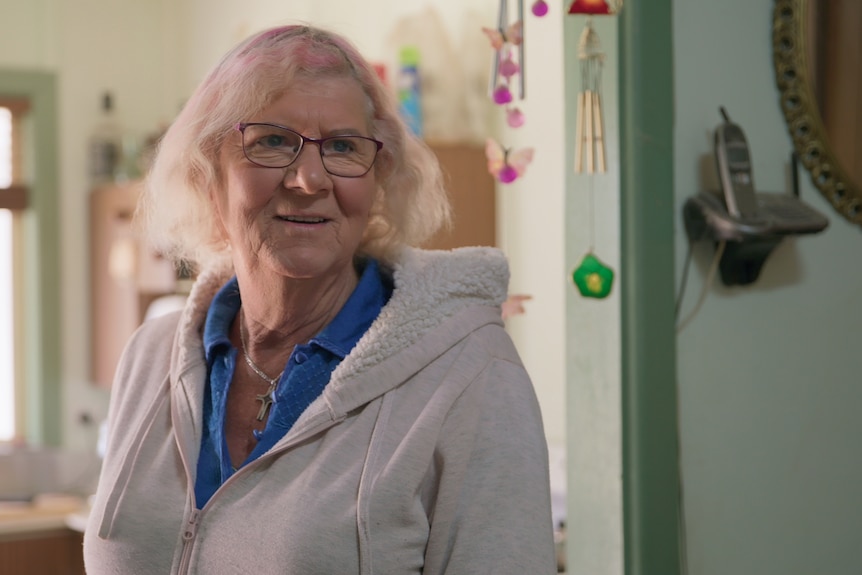 A woman stands in her house, wearing glasses and a grey jacket. She has light-coloured hair with pink throughout.