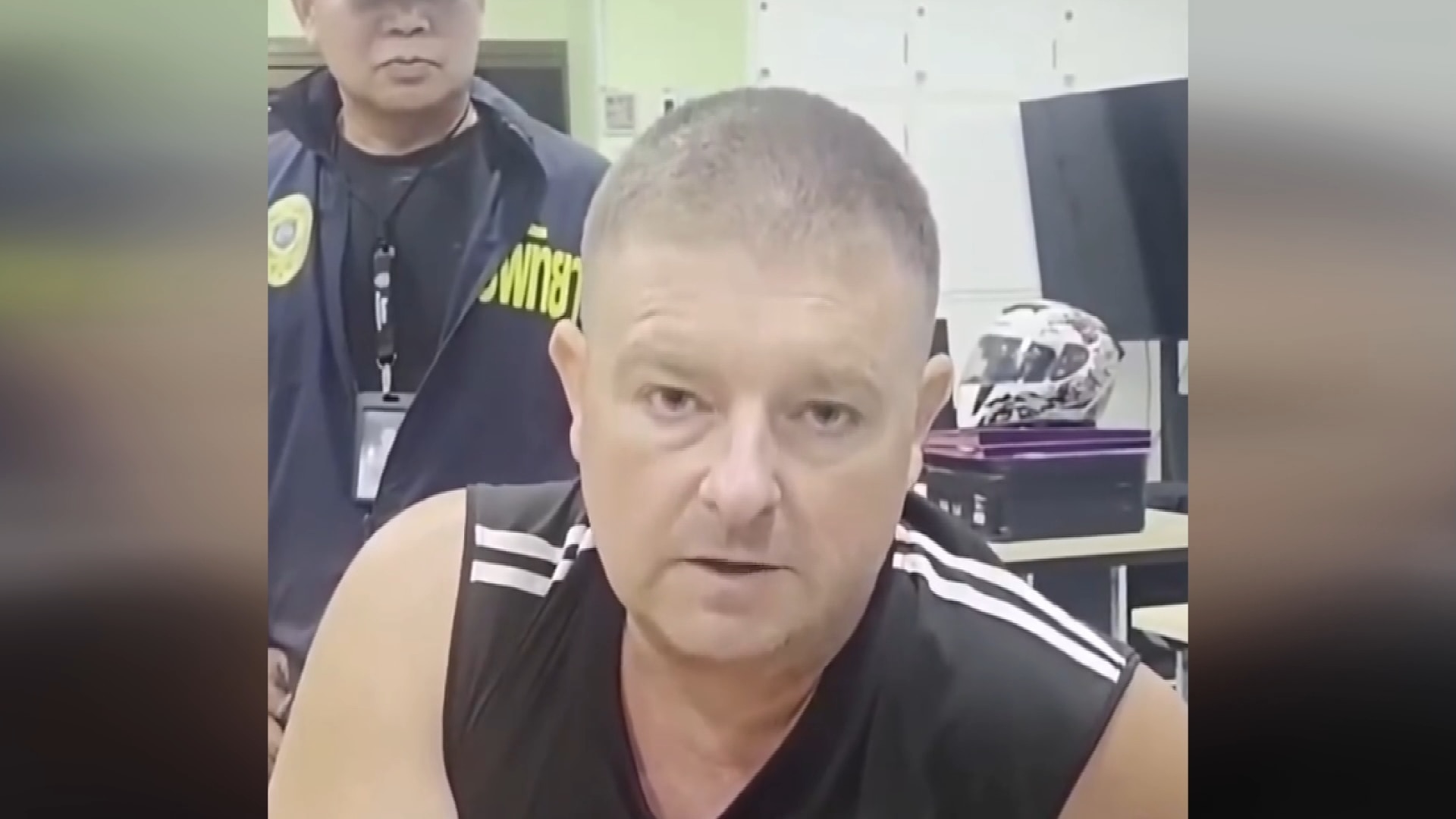 Mobile phone vision of a man speaking to the camera wearing a black singlet. A police officer stands behind him