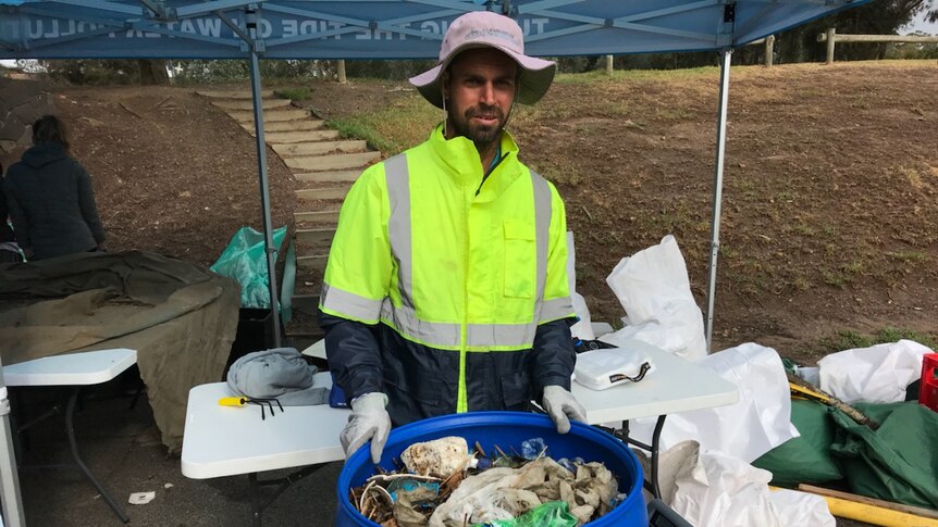 A man weighing a large blue bin filled with rubbish standing outside under a gazebo in a fluorescent yellow jacket and hat