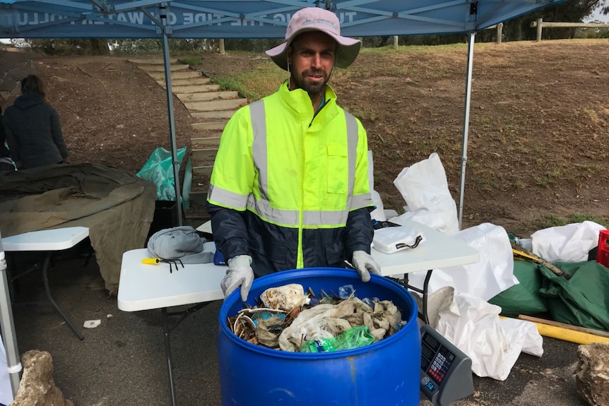 A man weighing a large blue bin filled with rubbish standing outside under a gazebo in a fluorescent yellow jacket and hat