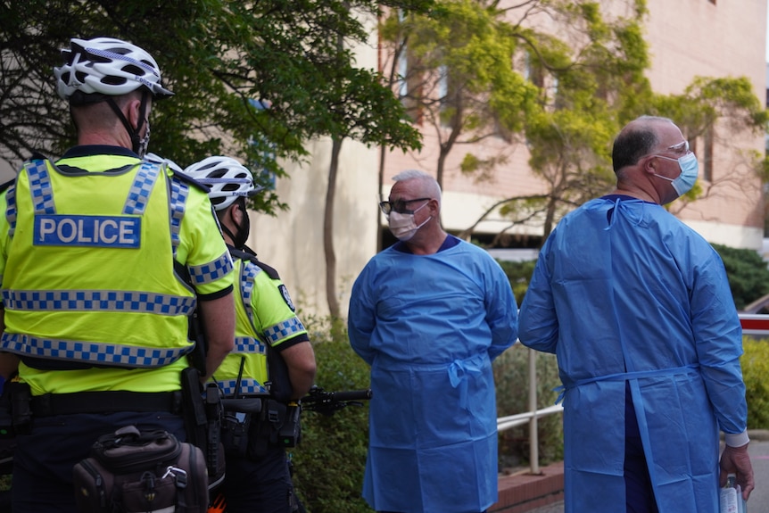 Two men in blue medical gowns and masks speak with two police officers.