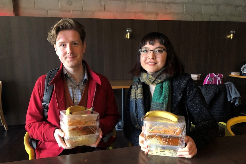 Ezgi and Stuart looks at the camera while holding parcels of food including bananas and pasta.