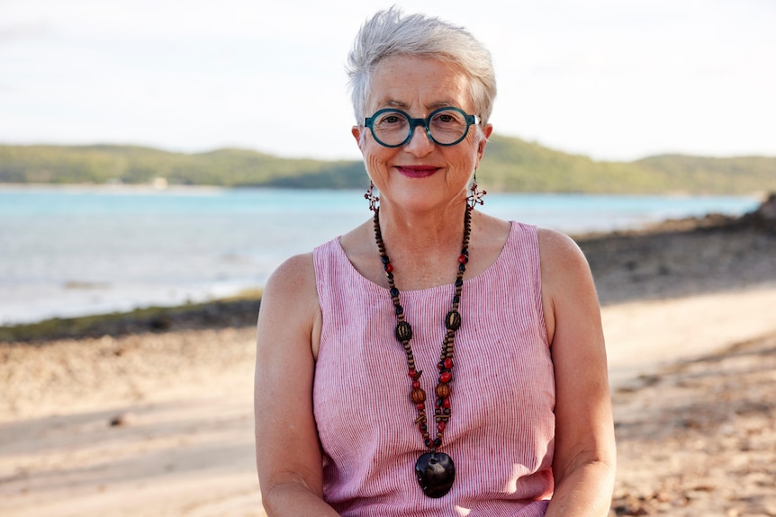 Ruth Stewart wears a pink top, beads and green glasses while standing on a beach.