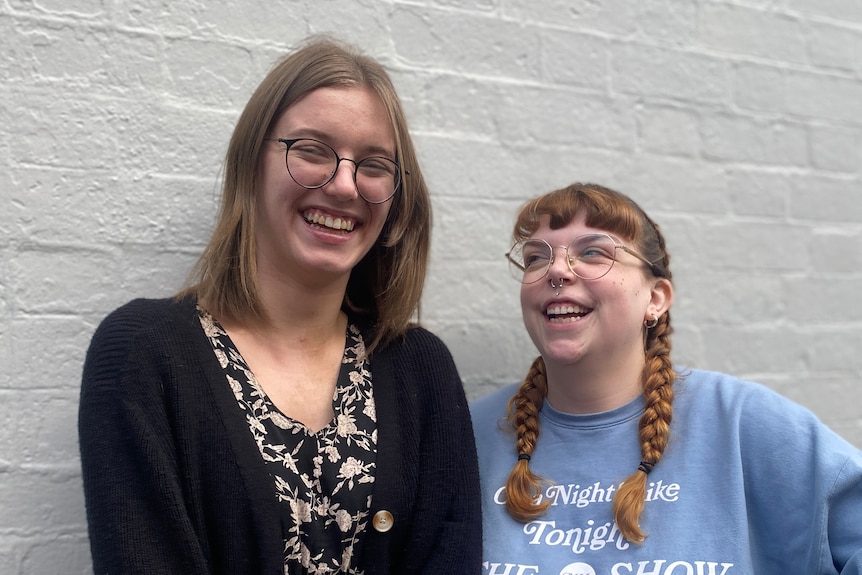 two young women stand next to one other smiling. Both wear reading glasses and one has her hair in two plaits. 