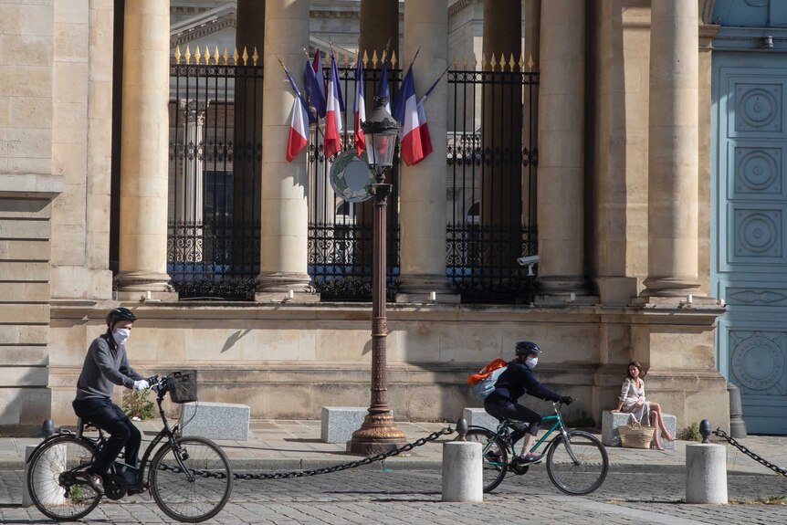 Two cyclists ride past a building with French blue, white and red flags hanging, with a woman sitting in the sun out front.