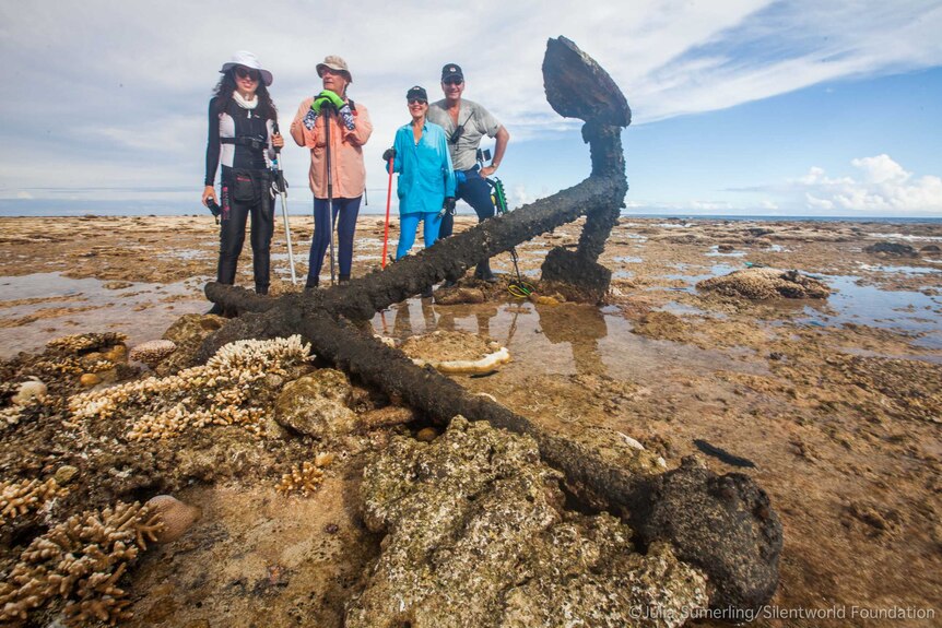 Four people stand behind a rusty anchor on top of a dry reef