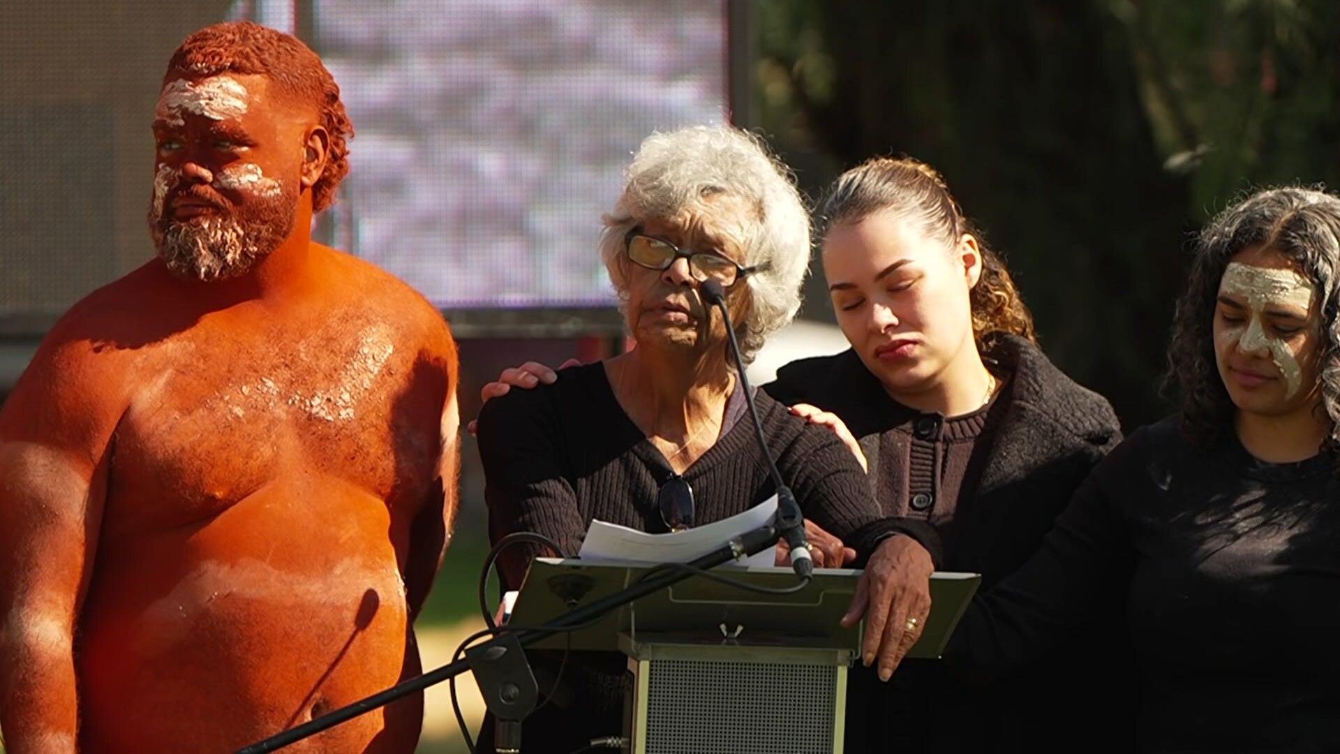 A woman surrounded by people speaks at a lectern.