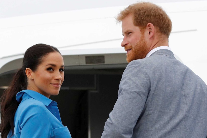 Woman with long, dark hair in blue collared dress holds hand of tall man with red hair and red beard wearing grey suit