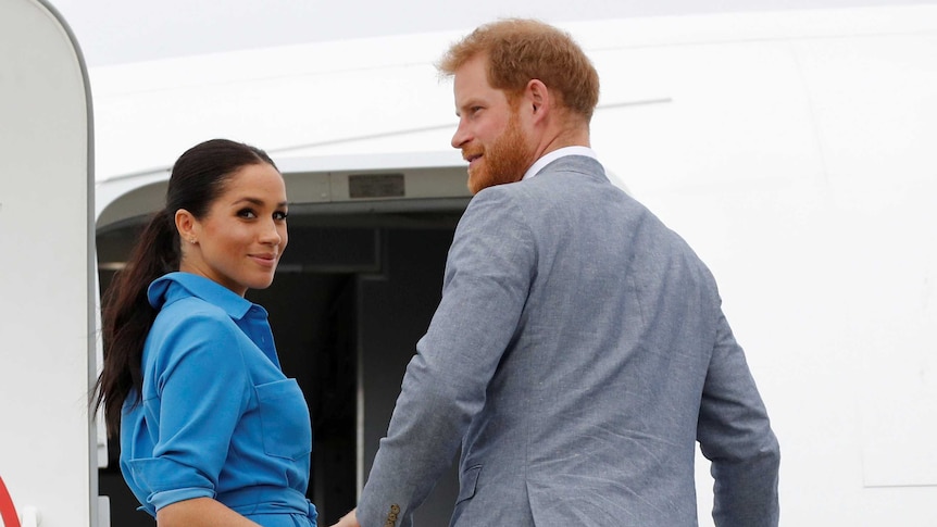 Woman with long, dark hair in blue collared dress holds hand of tall man with red hair and red beard wearing grey suit