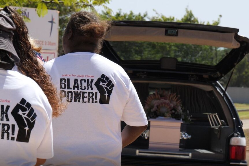 Two women stand near a hearse with a casket loaded in the back. They are wearing t-shirts reading 'Justice for Joyce'.
