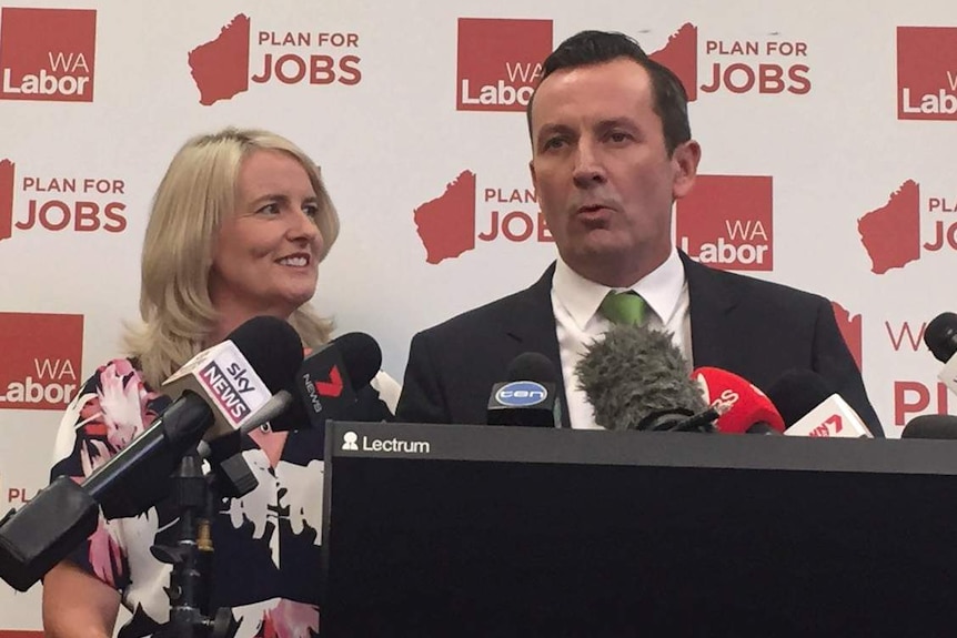 Mark McGowan stands at a podium talking into microphones with wife Sarah along side him.