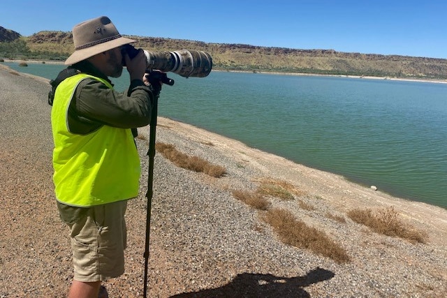 A man holds a camera set up on a stand next to a large pond.