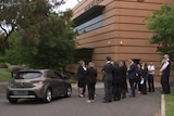 A grey hatchback outside an orange building with people standing around in suits