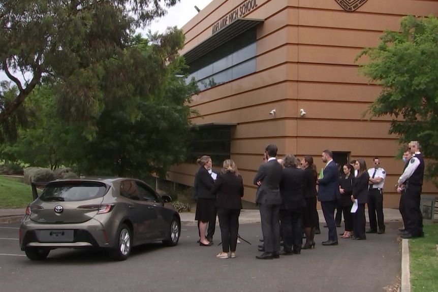 A grey hatchback outside an orange building with people standing around in suits