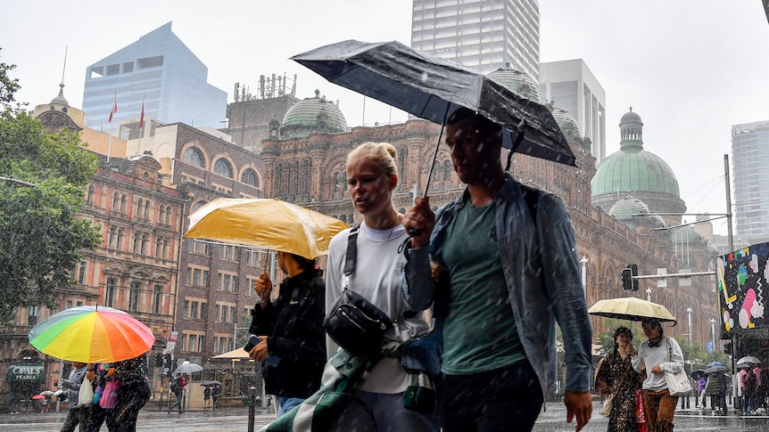 A man and woman share an umbrella as they walk down a wet Sydney street with large buildings behind them