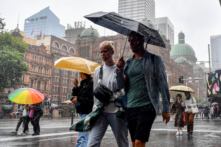 A man and woman share an umbrella as they walk down a wet Sydney street with large buildings behind them