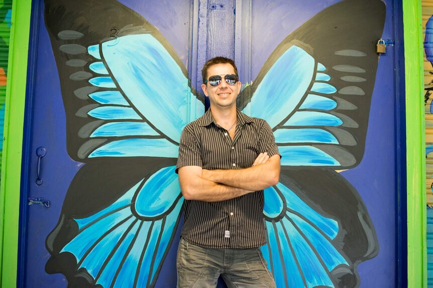 A man wearing sunglasses poses in front of a set of butterfly wings painted on a wall, making it look like the wings are his.