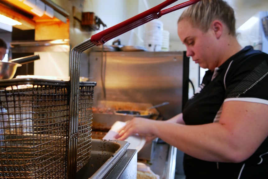 A deep frier is on in the foreground and a young woman is busy packing a takeaway order next to it