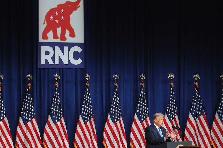 A man in a suit stands at a podium on stage with American flags in the background and a sign saying RNC.