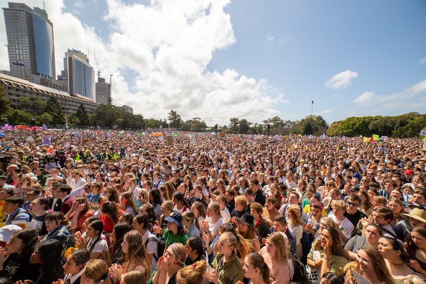 A crowd of tens of thousands of students looking up at a stage, it's sunny and people are in bright clothing with signs.