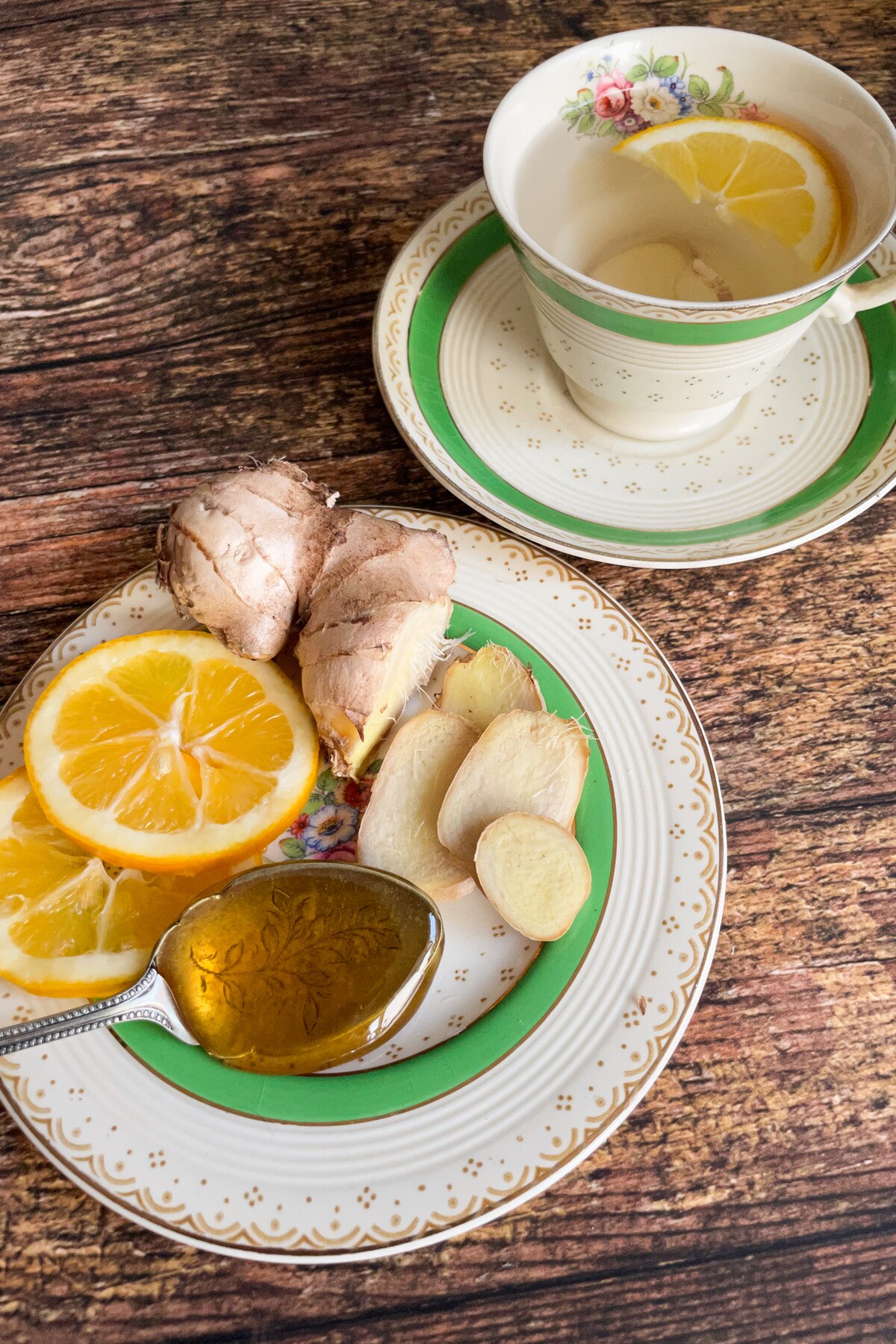 A tea cup with ginger and lemon slices in hot water next to a plate with ginger rot, lemon and honey displayed.