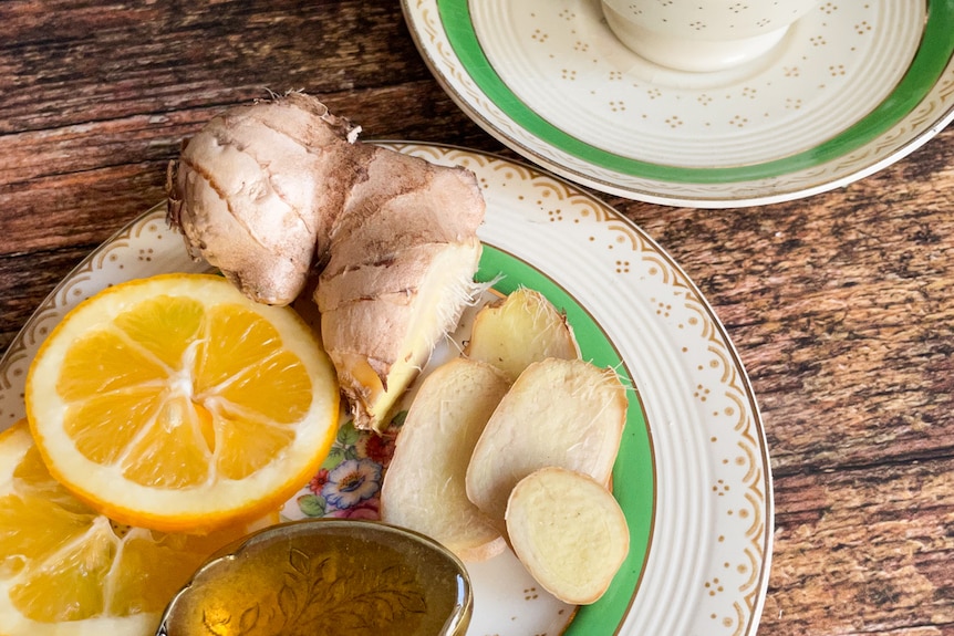 A tea cup with ginger and lemon slices in hot water next to a plate with ginger rot, lemon and honey displayed.