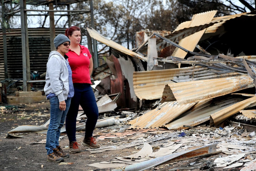 A woman in a red t-shirt and a woman in a beanie and cardigan stand among the ruins of a burnt house