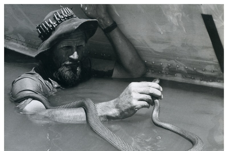 Harry Butler in the water next to a boat, holds a king brown snake's head above water.