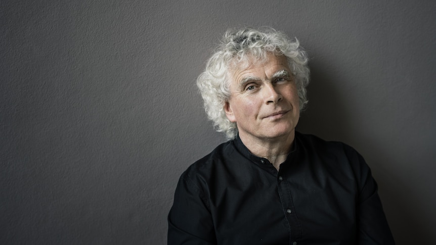 Simon Rattle against a grey wall looking at the camera
