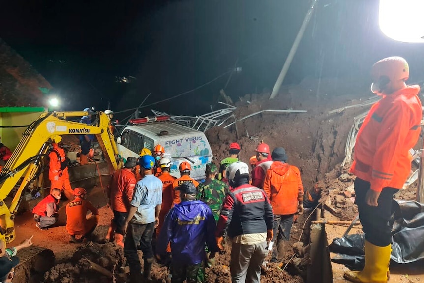 A night-time shot shows workers in orange jackets stand with excavating equipment in village rubble.